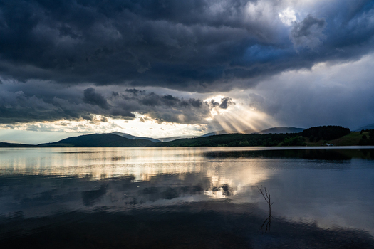 View of a calm lake reflecting dramatic storm clouds and sunbeams breaking through the sky over distant mountains in Sliven Bulgaria. View of a calm lake reflecting dramatic storm clouds and sunbeams breaking through the sky over distant mountains in Sliven Bulgaria.