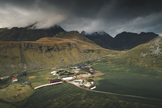 Aerial view of the rural landscape near Vik Strand featuring red farm buildings green agricultural fields and dramatic mountains under a moody overcast sky in Leknes Nordland Norway.