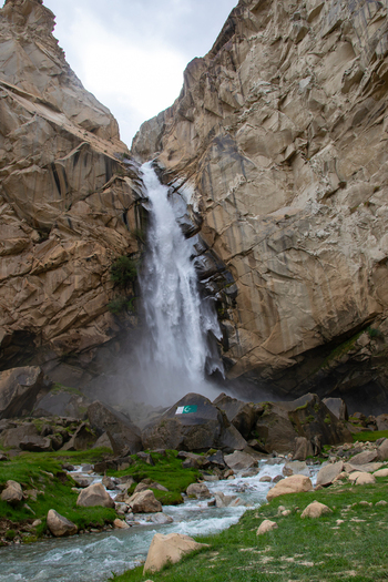View of Khamosh Waterfall cascading down steep rocky cliffs into a mountain stream with green grass and boulders in Skardu Gilgit Pakistan.