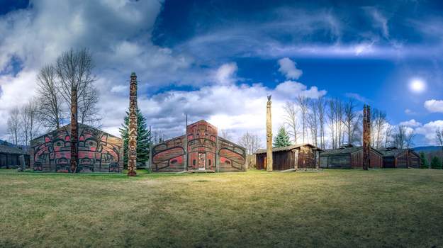 View of the traditional wooden longhouses and carved totem poles at Ksan Historical Village under a bright blue sky with scattered clouds in Ksan British Columbia Canada. View of the traditional wooden longhouses and carved totem poles at Ksan Historical Village under a bright blue sky with scattered clouds in Ksan British Columbia Canada.