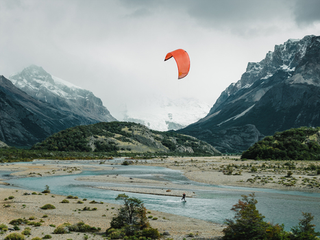 Aerial view of a kitesurfer on a river with an orange kite set against dramatic snow capped mountains and rocky terrain in El Chalten Santa Cruz Province Argentina. Aerial view of a kitesurfer on a river with an orange kite set against dramatic snow capped mountains and rocky terrain in El Chalten Santa Cruz Province Argentina.