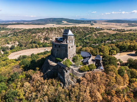 Aerial view of Holloko Castle a medieval stone fortress above forested hills dry farmland and scattered rural homes in Holloko Hungary on a clear late summer day. Aerial view of Holloko Castle a medieval stone fortress above forested hills dry farmland and scattered rural homes in Holloko Hungary on a clear late summer day.