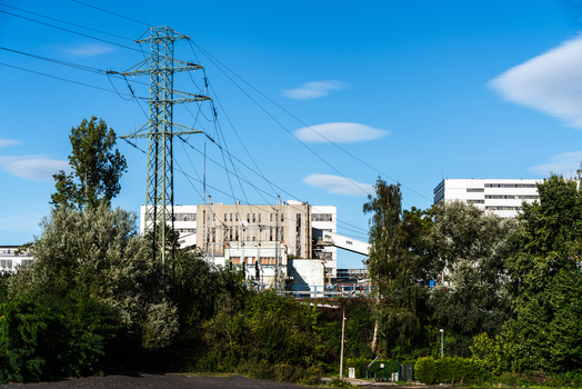 View of a metal power transmission tower standing near an industrial factory complex surrounded by lush green trees under a clear blue sky in Poland.