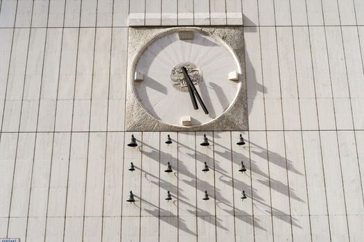 View of a large stone clock and a set of small bells casting shadows on a modern stone facade in Bratislava Bratislava Region Slovakia. View of a large stone clock and a set of small bells casting shadows on a modern stone facade in Bratislava Bratislava Region Slovakia.