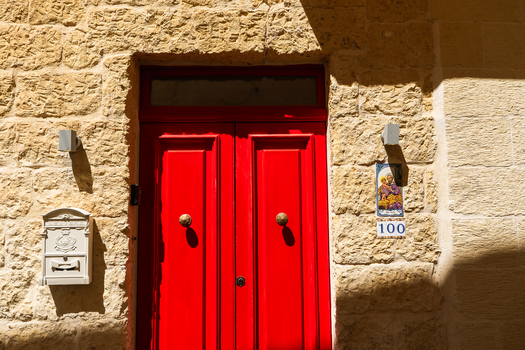 View of a traditional bright red wooden door set in a textured limestone wall with a white mailbox and religious tile in Zebbug Gozo Region Malta. View of a traditional bright red wooden door set in a textured limestone wall with a white mailbox and religious tile in Zebbug Gozo Region Malta.