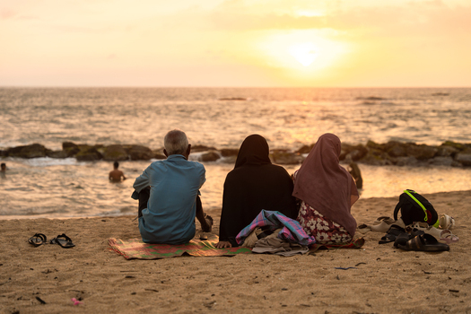 Sri Lanka 14 January 2026 View of three people sitting on a sandy beach watching the sunset over the Indian Ocean with rocky breakwaters and swimmers in the distance.