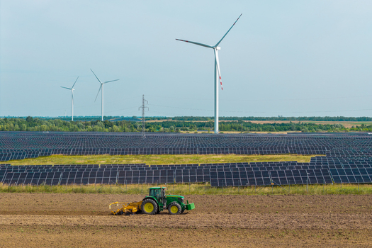 Aerial view of a large solar farm with rows of photovoltaic panels and wind turbines in the background with a green tractor working in a field in Poland.