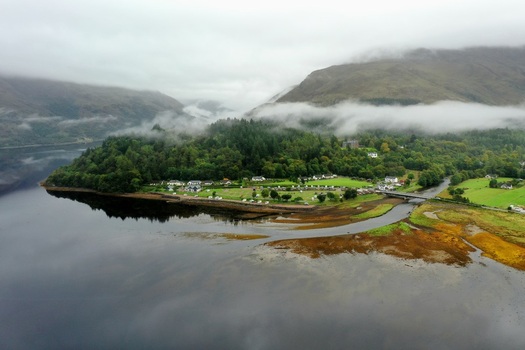 Aerial view of Glencoe featuring lush green forests a calm lake a small village with houses and misty mountains under a cloudy sky in Glencoe Scotland United Kingdom.