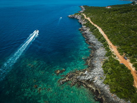 Aerial view of the rocky coastline of Aquarium Beach with turquoise waters a boat leaving a wake and a dirt road winding through lush green hills in Jala Vlore County Albania. Aerial view of the rocky coastline of Aquarium Beach with turquoise waters a boat leaving a wake and a dirt road winding through lush green hills in Jala Vlore County Albania.