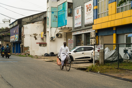 Sri Lanka 15 February 2026 View of a man in traditional white clothing riding a bicycle along a street lined with commercial buildings and parked vehicles.