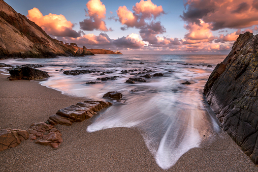 View of a rocky beach in Howth with long exposure waves washing over the sand under pink tinted clouds at sunset in Ireland. View of a rocky beach in Howth with long exposure waves washing over the sand under pink tinted clouds at sunset in Ireland.