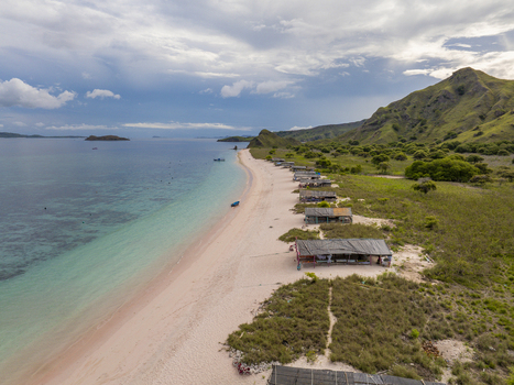 Aerial view of Komodo National Park featuring a pink sand beach with wooden huts turquoise waters and green hills in Komodo East Nusa Tenggara Indonesia. Aerial view of Komodo National Park featuring a pink sand beach with wooden huts turquoise waters and green hills in Komodo East Nusa Tenggara Indonesia.