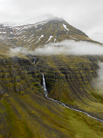 Aerial view of a waterfall cascading down a steep mossy mountain slope with snow patches and low clouds in the Eastern Region Iceland. Aerial view of a waterfall cascading down a steep mossy mountain slope with snow patches and low clouds in the Eastern Region Iceland.