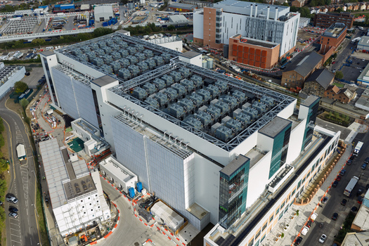 Aerial view of a large modern data center with industrial cooling units on the roof surrounded by urban infrastructure and roads in the United Kingdom. Aerial view of a large modern data center with industrial cooling units on the roof surrounded by urban infrastructure and roads in the United Kingdom.