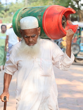 Cox s Bazar Chittagong Division Bangladesh 19 July 2026 View of an elderly man with a white beard carrying a large green and red gas cylinder on his shoulder while walking through the Rohingya refugee camp.