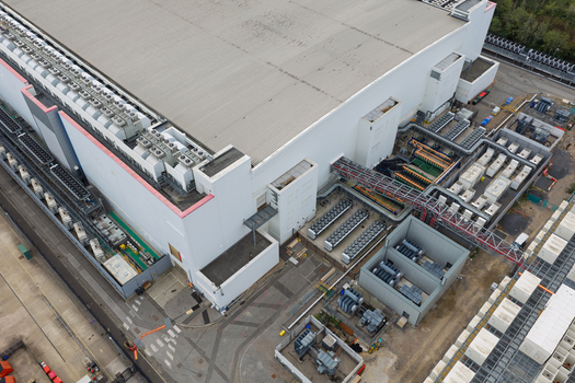 Aerial view of a large data center facility with a massive industrial building cooling units on the roof and electrical infrastructure in the surrounding yard United Kingdom.
