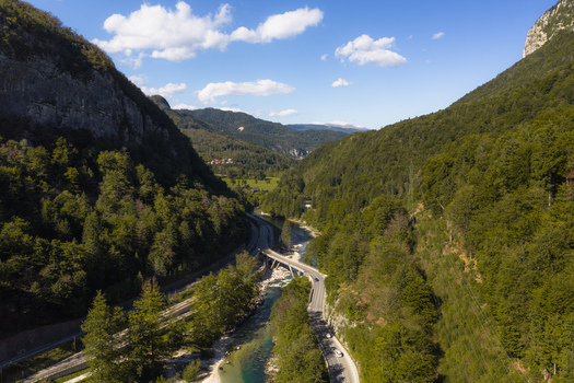 Aerial view of Sava Bohinjka river flowing through a lush green valley with a road bridge and forested mountain slopes under a clear blue sky Radovljica Slovenia.