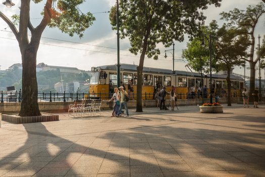 View of a yellow tram with people walking along the paved promenade lined with trees in Budapest Hungary.