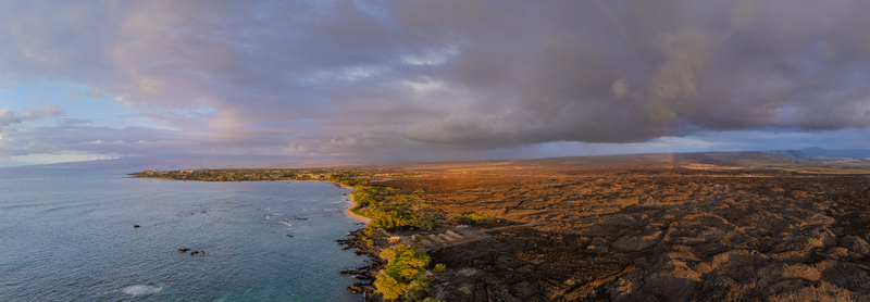 Aerial view of Waikoloa Beach with dark volcanic lava fields turquoise ocean waters and a faint rainbow under dramatic storm clouds in Waikoloa Village Hawaii United States.