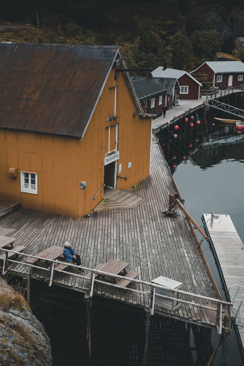 Nusfjord Nordland Norway 07 September 2022 View of the historic Nusfjord fishing village with a person on a pier beside a yellow rorbu surrounded by calm water and red huts. Nusfjord Nordland Norway 07 September 2022 View of the historic Nusfjord fishing village with a person on a pier beside a yellow rorbu surrounded by calm water and red huts.