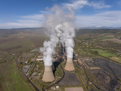 Aerial view of a large coal fired power plant with massive cooling towers emitting thick white steam plumes into a blue sky surrounded by industrial grounds and rural landscape in Bulgaria.