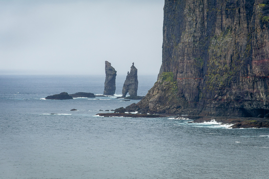 View of the dramatic sea cliffs and the Risin og Kellingin sea stacks rising from the ocean under a cloudy sky in the Faroe Islands.
