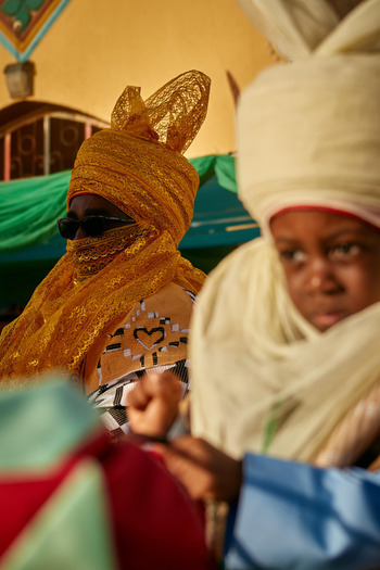Zaria city Nigeria 22 March 2026 View of a regal figure in a golden textured turban and dark shades alongside a young boy in soft cream attire amidst vibrant fabrics. Zaria city Nigeria 22 March 2026 View of a regal figure in a golden textured turban and dark shades alongside a young boy in soft cream attire amidst vibrant fabrics.