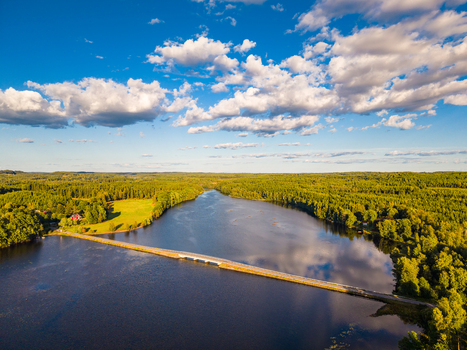 Aerial view of a bridge crossing a calm lake surrounded by lush green forests and meadows under a bright blue sky with fluffy clouds in Gallared Halland County Sweden. Aerial view of a bridge crossing a calm lake surrounded by lush green forests and meadows under a bright blue sky with fluffy clouds in Gallared Halland County Sweden.