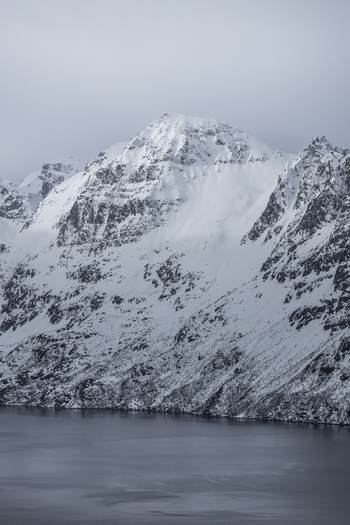 View of a snow covered mountain peak rising above the calm waters of a fjord under an overcast sky Ersfjord Troms Norway. View of a snow covered mountain peak rising above the calm waters of a fjord under an overcast sky Ersfjord Troms Norway.