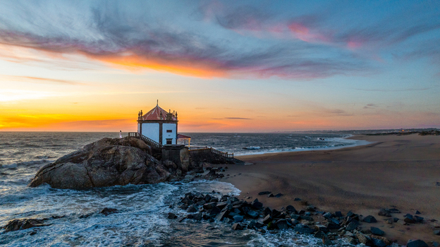 Aerial view of the Capela do Senhor da Pedra chapel built on a rocky outcrop at sunset with waves crashing on the beach in Miramar Porto Portugal.