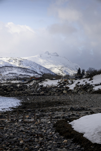 View of the rocky shoreline and snow covered mountains under a cloudy sky in Kvaloya Troms Norway. View of the rocky shoreline and snow covered mountains under a cloudy sky in Kvaloya Troms Norway.