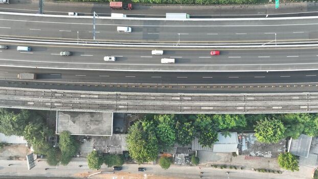Aerial view of a multi lane highway with cars and trucks adjacent to a railway line and lush green trees in Bekasi Barat West Java Indonesia. Aerial view of a multi lane highway with cars and trucks adjacent to a railway line and lush green trees in Bekasi Barat West Java Indonesia.