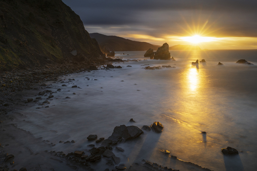View of the rocky coastline of Gaztelugatxeko Doniene with a golden sunset starburst over the ocean and misty waves Gaztelugatxeko Doniene Euskadi Spain. View of the rocky coastline of Gaztelugatxeko Doniene with a golden sunset starburst over the ocean and misty waves Gaztelugatxeko Doniene Euskadi Spain.
