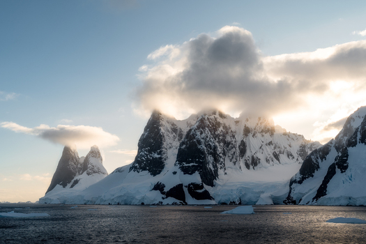 View of snow covered mountain peaks with clouds resting on the summits and icebergs floating in the cold sea during the golden hour of sunset Antarctica.