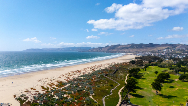Aerial view of the sandy coastline with dunes ocean waves and rolling hills under a blue sky in Morro Bay California United States.