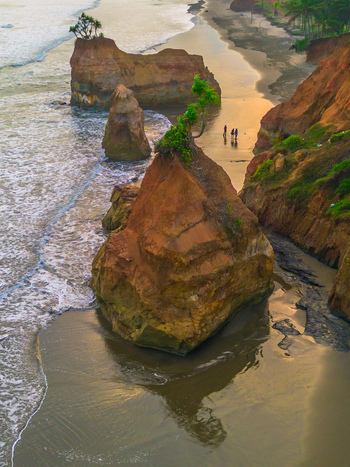 Aerial view of dramatic coastal rock formations with lush vegetation on top surrounded by dark sand beach and gentle waves at sunset Bali Indonesia. Aerial view of dramatic coastal rock formations with lush vegetation on top surrounded by dark sand beach and gentle waves at sunset Bali Indonesia.