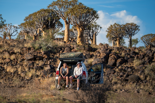 Aussenkehr Namibia 06 March 2026 View of two people sitting on the back of a vehicle enjoying the arid landscape dotted with quiver trees under a clear blue sky.. Aussenkehr Namibia 06 March 2026 View of two people sitting on the back of a vehicle enjoying the arid landscape dotted with quiver trees under a clear blue sky..