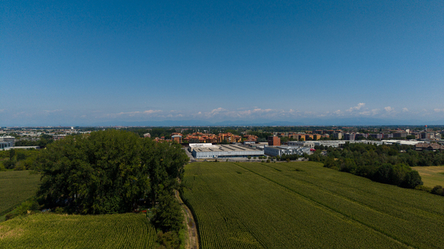 Aerial view of lush green cornfields and industrial buildings with a residential area in the distance under a clear blue sky in Milan Lombardy Italy. Aerial view of lush green cornfields and industrial buildings with a residential area in the distance under a clear blue sky in Milan Lombardy Italy.