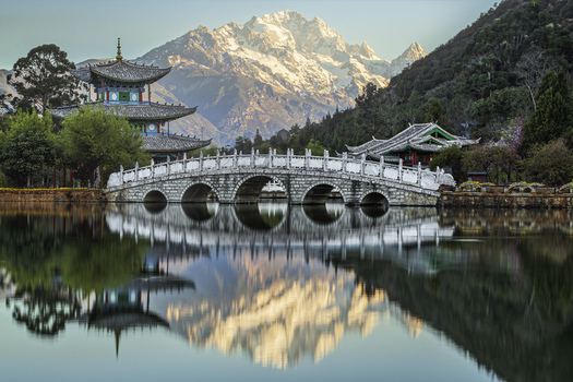 Aerial view of the iconic Suocui Bridge at Black Dragon Pool Park with the snow capped Jade Dragon Snow Mountain reflecting in the calm water in Lijiang Yunnan China.