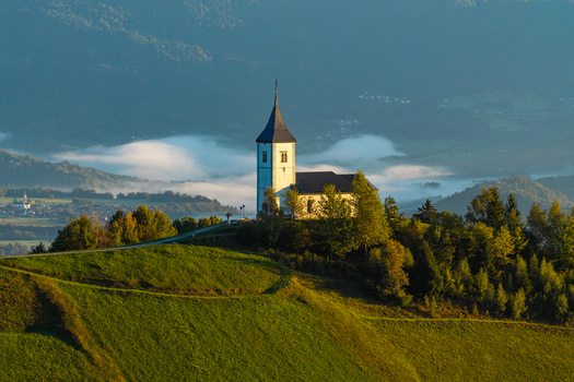 Aerial view of the Church of Saints Primus and Felician in Jamnik surrounded by lush green hills and morning mist in the valley Kranj Slovenia. Aerial view of the Church of Saints Primus and Felician in Jamnik surrounded by lush green hills and morning mist in the valley Kranj Slovenia.