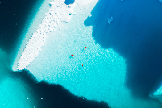 Aerial view of three people swimming in crystal clear turquoise water next to a massive white iceberg Greenland. Aerial view of three people swimming in crystal clear turquoise water next to a massive white iceberg Greenland.