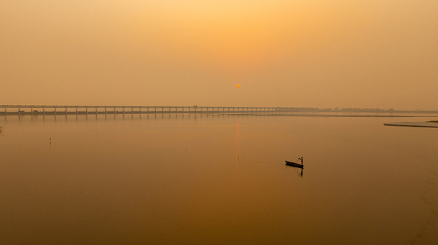 Aerial view of a lone fisherman in a small wooden boat on a calm river at sunset with a long bridge spanning the horizon in Nepal.