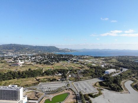 Aerial view of the coastal landscape featuring a large hotel complex expansive parking areas lush green fields and the blue ocean in the distance in Motobu Okinawa Japan. Aerial view of the coastal landscape featuring a large hotel complex expansive parking areas lush green fields and the blue ocean in the distance in Motobu Okinawa Japan.