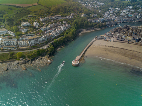 Aerial view of the coastal town of Looe featuring a small boat navigating the harbour entrance the sandy beach and hillside residential buildings in Looe Cornwall United Kingdom. Aerial view of the coastal town of Looe featuring a small boat navigating the harbour entrance the sandy beach and hillside residential buildings in Looe Cornwall United Kingdom.