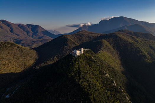 Aerial view of Santuario del Santissimo Salvatore perched on a mountain peak surrounded by lush forested hills and distant peaks under a clear blue sky Montella Campania Italy. Aerial view of Santuario del Santissimo Salvatore perched on a mountain peak surrounded by lush forested hills and distant peaks under a clear blue sky Montella Campania Italy.