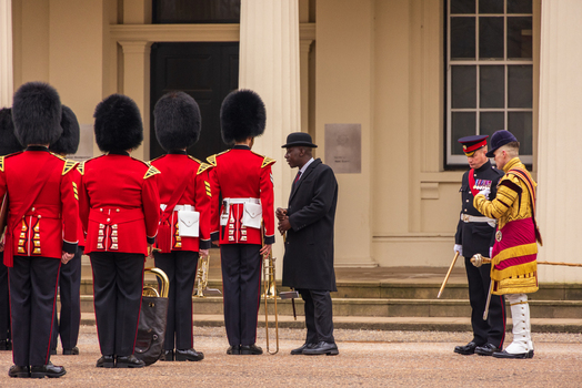 London United Kingdom 12 May 2025 View of the King s Guard in red tunics and bearskin hats standing outside a royal palace with officers and a man in a bowler hat.