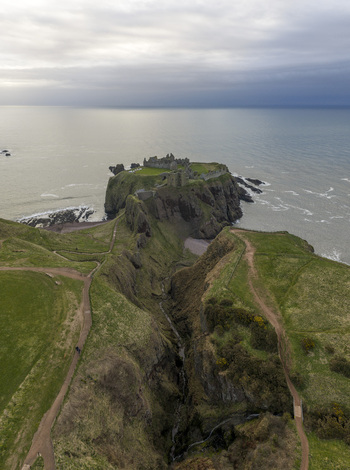 Aerial view of Dunnottar Castle perched on a rocky headland overlooking the North Sea with green cliffs and walking paths in Stonehaven Scotland United Kingdom. Aerial view of Dunnottar Castle perched on a rocky headland overlooking the North Sea with green cliffs and walking paths in Stonehaven Scotland United Kingdom.