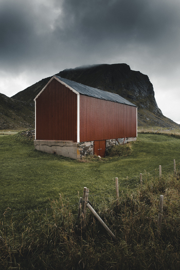 View of a red barn with a stone foundation set in a grassy field near Haukland Beach with a dramatic mountain in the background in Leknes Nordland Norway. View of a red barn with a stone foundation set in a grassy field near Haukland Beach with a dramatic mountain in the background in Leknes Nordland Norway.