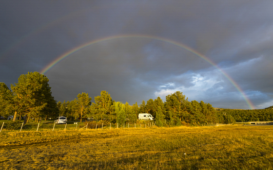 View of a vibrant rainbow arcs across a golden field meeting lush green trees under a dramatic sky with campervans nestled in nature Alta Finnmark Norway. View of a vibrant rainbow arcs across a golden field meeting lush green trees under a dramatic sky with campervans nestled in nature Alta Finnmark Norway.