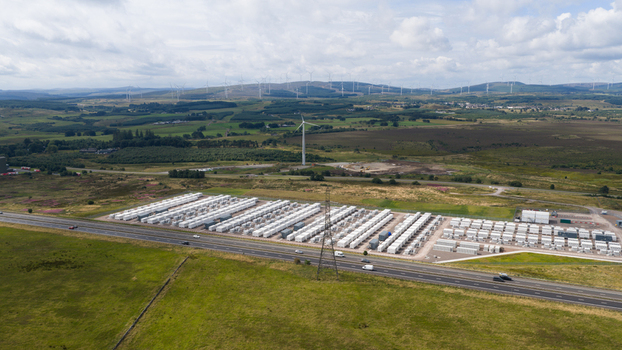 Aerial view of a large battery energy storage system with rows of white containers next to a highway in a rural landscape with wind turbines in Coalburn Scotland United Kingdom.